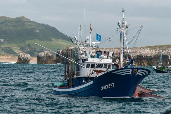 Barco de pesca artesanal faenando al curricán en el mar Cantábrico, varias cañas largas asomando por la popa, día soleado