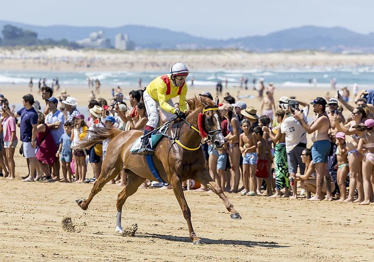 Carrera de caballos sobre la arena durante el Derby de Loredo, con público en la orilla