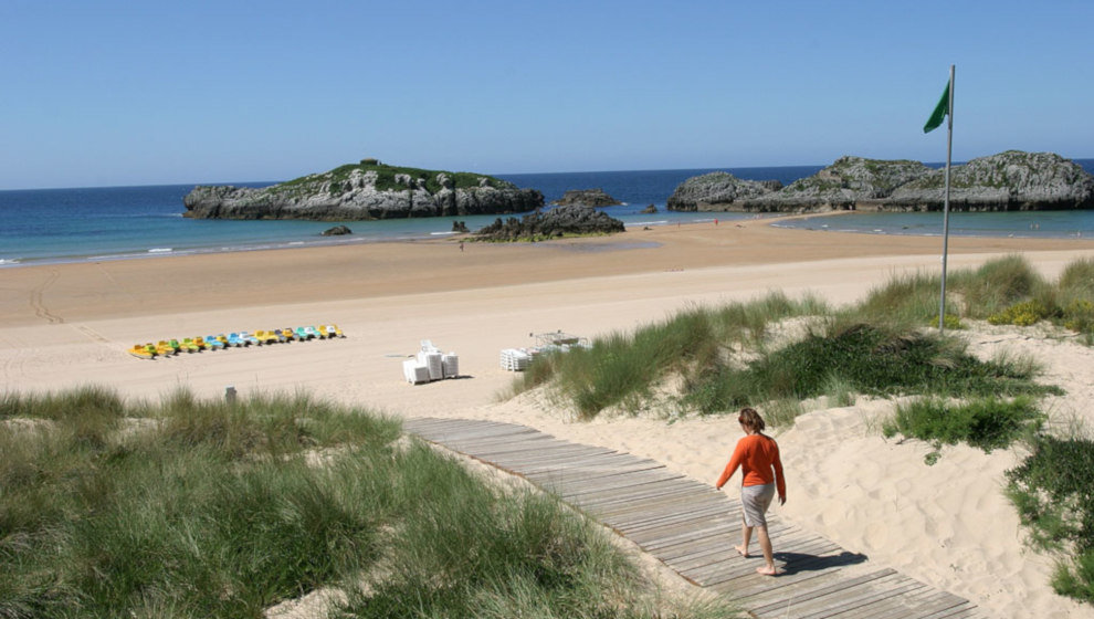Vista general de una playa cerca de Bareyo con la marea baja y el mástil de banderas vacío en un día soleado, sin bañistas destacados en primer plano.