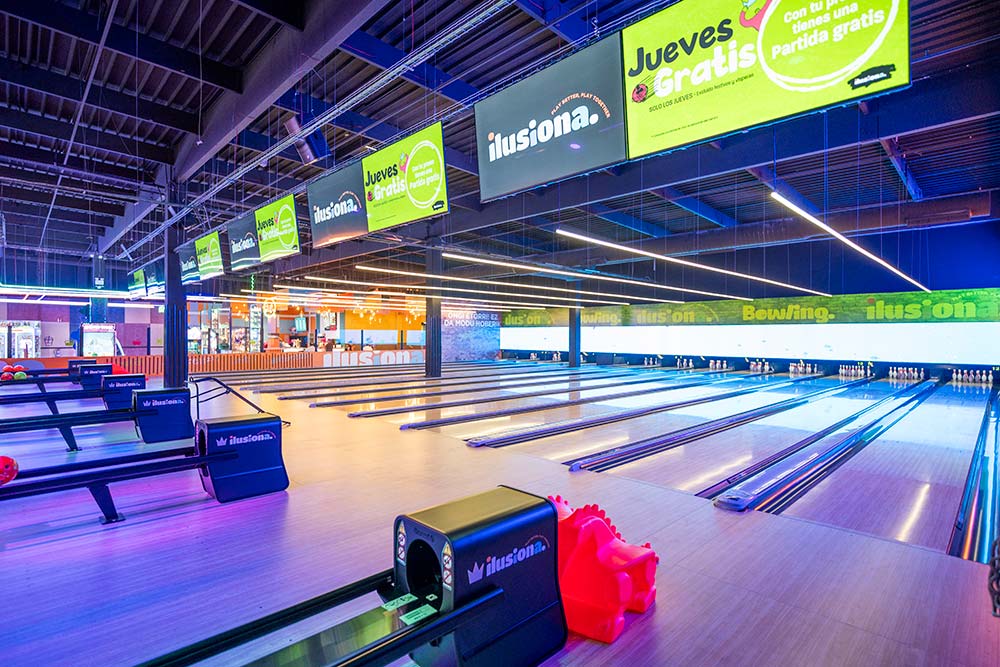 Pista de bolos interior iluminada con bolos en pie al fondo, ambiente de centro comercial.