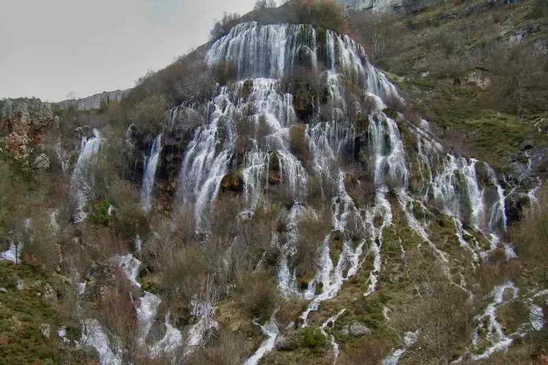 Pared rocosa de toba con agua emanando en un salto de unos 100 metros de altura sobre un valle fluvial (Cascada del Tobazo, Valderredible).