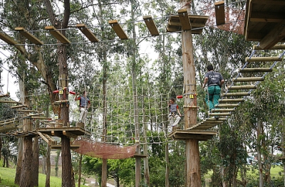 Persona con arnés y casco cruzando un puente colgante entre árboles en el circuito Mixto de Tirolinas Go! Santander (antes Forestal Park), día soleado.