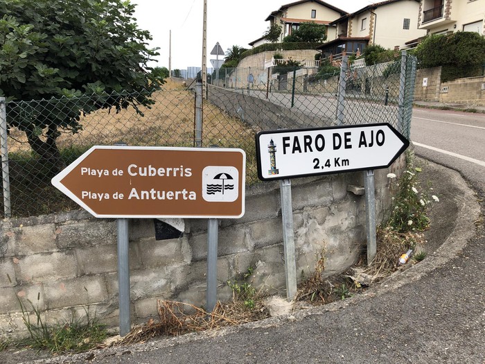 Carretera rural de acceso al barrio de Ajo, Bareyo, con campo verde y señalización local a los lados
