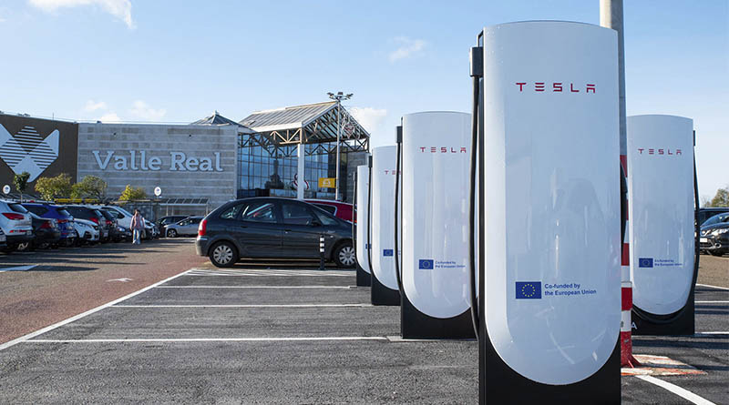 Coche eléctrico cargando en un punto Tesla Supercharger junto a un centro comercial